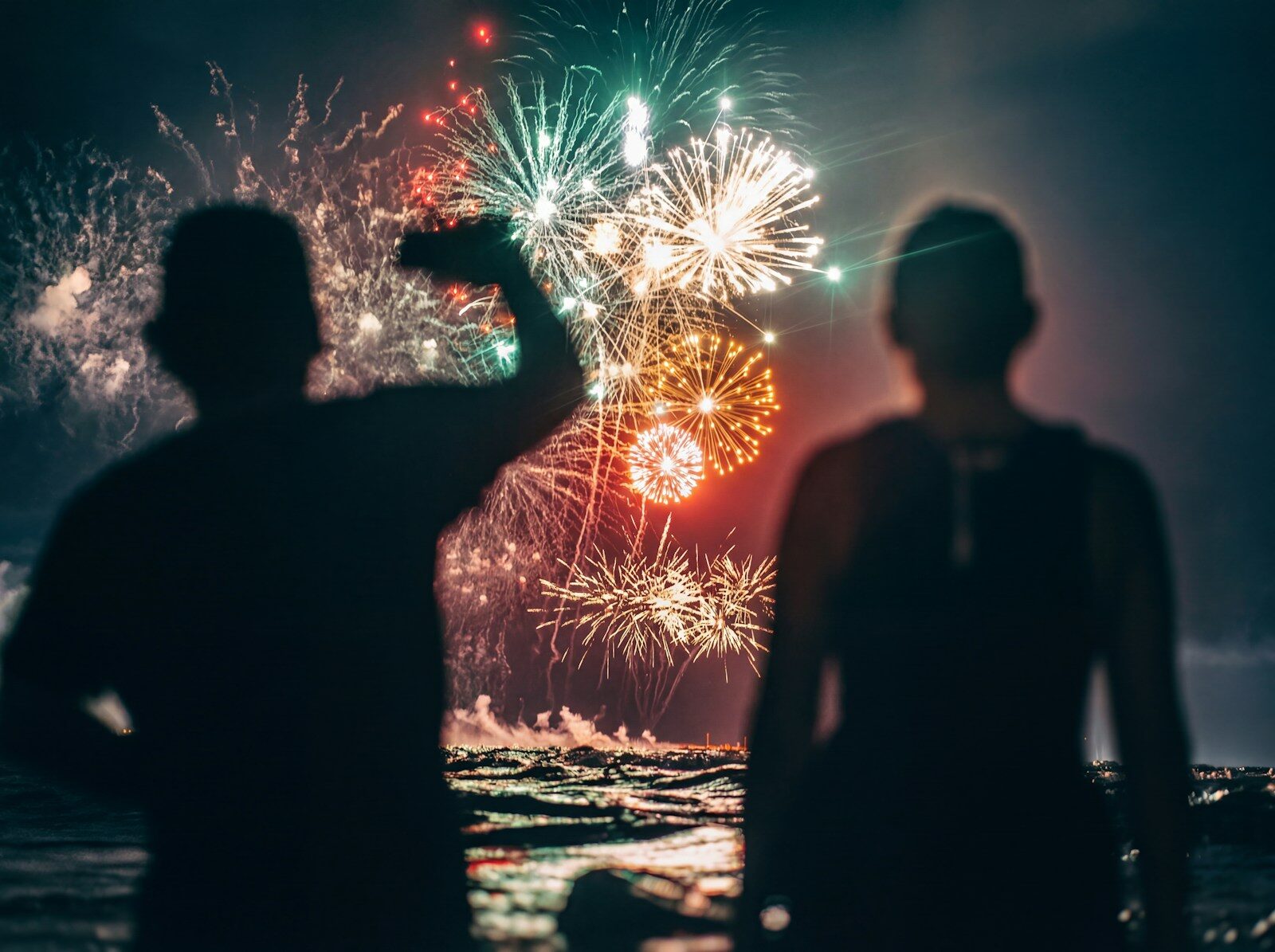Silhouetted figures watching fireworks display over water.